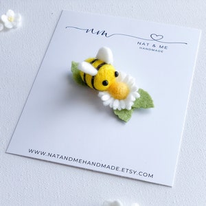 May include: A felt bee with black stripes and yellow body is perched on a white felt daisy with green leaves. The bee has black eyes and a yellow face. The bee and daisy are on a white card with the text "Nat & Me Handmade" and a website address.