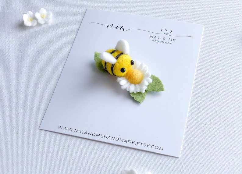 May include: A felt bee with black stripes and yellow body is perched on a white felt daisy with green leaves. The bee has black eyes and a yellow face. The bee and daisy are on a white card with the text "Nat & Me Handmade" and a website address.