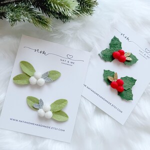 May include: Two sets of handmade Christmas hair clips. One set features green felt leaves with white pom-poms and silver glitter accents. The other set has green felt holly leaves with red pom-poms and gold glitter accents. The cards read "NAT & ME" and "www.natandmehandmade.etsy.com".