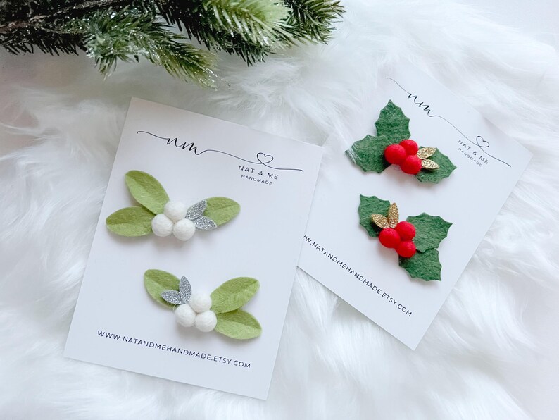 May include: Two sets of handmade Christmas hair clips. One set features green felt leaves with white pom-poms and silver glitter accents. The other set has green felt holly leaves with red pom-poms and gold glitter accents. The cards read "NAT & ME" and "www.natandmehandmade.etsy.com".