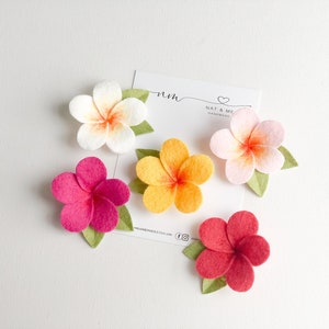 May include: Five felt flower embellishments in various colours, including pink, red, white, and yellow. The flowers are arranged on a white surface with a card that reads "Nat & Me Handmade".