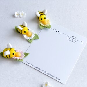 May include: Three felt bumblebees with black and yellow stripes are perched on white and yellow flowers. The bees are handmade and have black eyes. The flowers have green leaves. The bees are on a white background with a white card that says "Nat & Me Handmade" and "www.natandmehandmade.etsy.com".