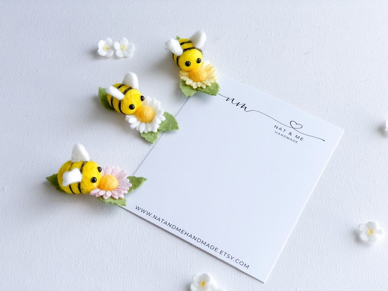 May include: Three felt bumblebees with black and yellow stripes are perched on white and yellow flowers. The bees are handmade and have black eyes. The flowers have green leaves. The bees are on a white background with a white card that says "Nat & Me Handmade" and "www.natandmehandmade.etsy.com".