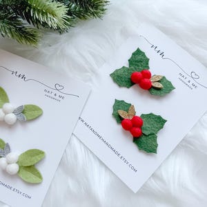 May include: Two sets of Christmas hair clips. One set features green felt leaves with white berries and silver glitter accents. The other set has green felt leaves with red berries and gold glitter accents. Each set is on a white card with the text "NAT & ME HANDMADE".
