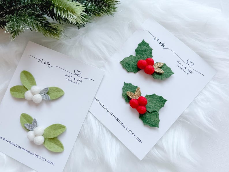 May include: Two sets of Christmas hair clips. One set features green felt leaves with white berries and silver glitter accents. The other set has green felt leaves with red berries and gold glitter accents. Each set is on a white card with the text "NAT & ME HANDMADE".