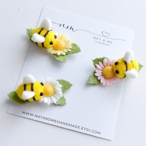 May include: Three felt bee and flower hair clips. The bees are yellow with black stripes and white wings. The flowers are yellow, pink, and white. The clips are handmade by Nat & Me.