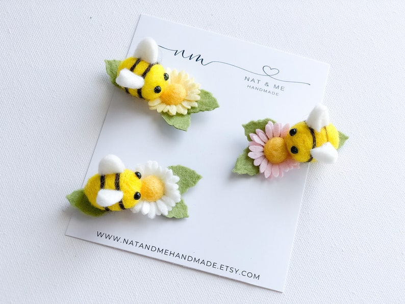 May include: Three felt bee and flower hair clips. The bees are yellow with black stripes and white wings. The flowers are yellow, pink, and white. The clips are handmade by Nat & Me.