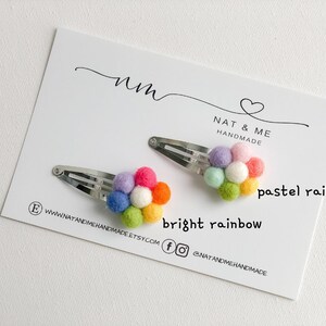 K&ouml;nnte beinhalten: Zwei regenbogenfarbene Filzblumen-Haarklammern. Eine Klammer hat einen Pastellregenbogen und die andere einen leuchtenden Regenbogen. Die Klammern befinden sich auf einer wei&szlig;en Karte mit dem Text "Nat & Me Handmade" und "pastel rainbow" und "bright rainbow".