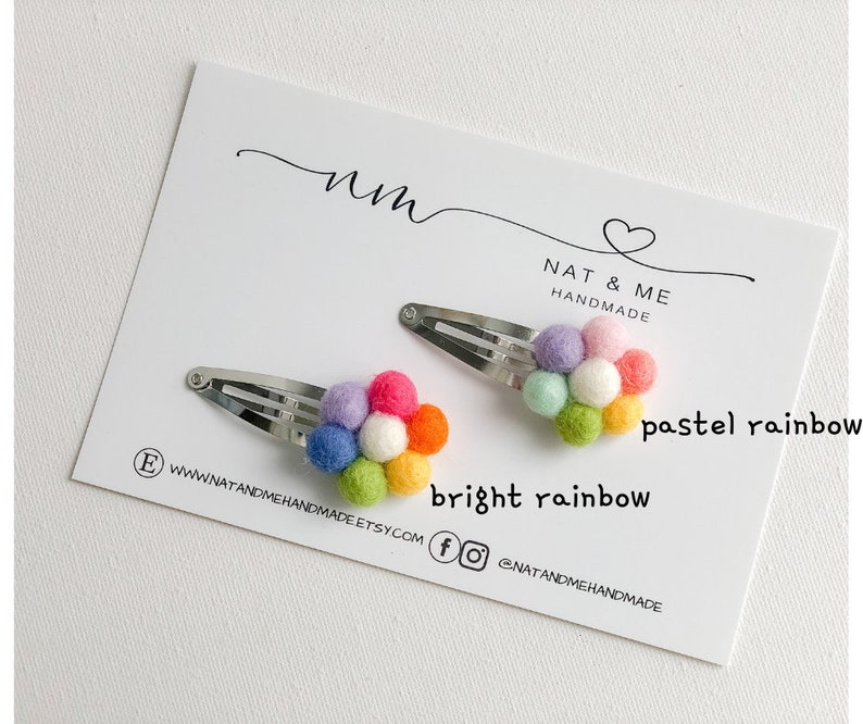 K&ouml;nnte beinhalten: Zwei regenbogenfarbene Filzblumen-Haarklammern. Eine Klammer hat einen Pastellregenbogen und die andere einen leuchtenden Regenbogen. Die Klammern befinden sich auf einer wei&szlig;en Karte mit dem Text "Nat & Me Handmade" und "pastel rainbow" und "bright rainbow".