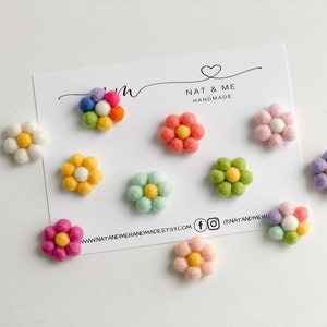 K&ouml;nnte beinhalten: Sortiment an Filzblumen-Verzierungen in verschiedenen Farben, darunter Rosa, Gelb, Gr&uuml;n, Wei&szlig; und Lila. Die Blumen sind auf einer wei&szlig;en Karte mit dem Text "Nat & Me Handmade" angeordnet.