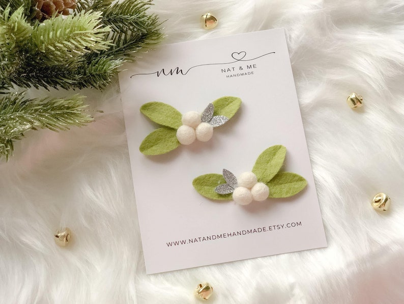 May include: Two handmade mistletoe hair clips. Each clip features light green felt leaves, white pom-pom berries, and silver glitter accents. The clips are displayed on a white card with the text "NAT & ME HANDMADE".