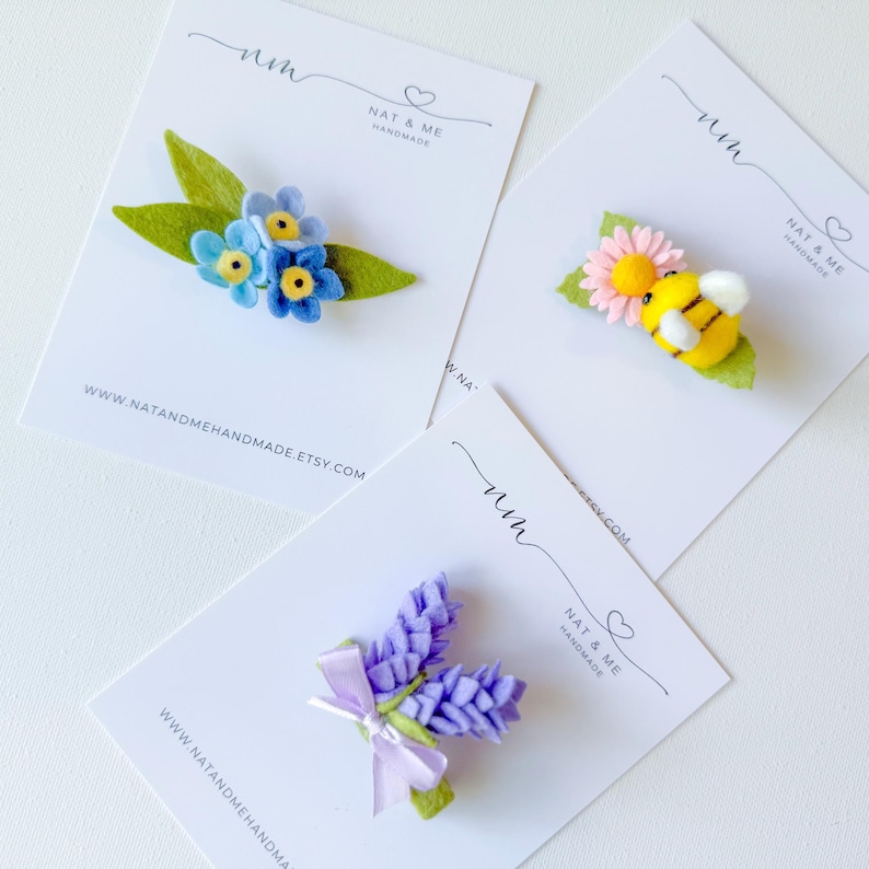 May include: Three felt hair clips on white display cards. One features blue flowers with green leaves, another a yellow bee on a pink flower with a green leaf, and the third a purple lavender sprig with a light purple ribbon bow.