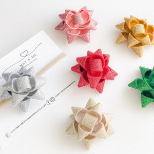 May include: A collection of handmade glitter bow hair accessories in various colors, including pink, red, gold, and green. One bow is attached to a beige headband. The bows are arranged on a white surface.