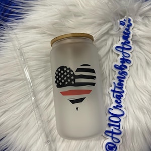 May include: A frosted glass tumbler with a bamboo lid features a red, white, and blue American flag heart design with a thin red line across the center. The tumbler is on a white fluffy surface with a blue and white sticker that says "@At10CreationsbyAtraiu".