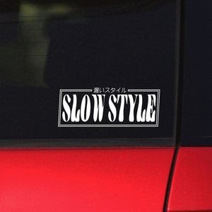 以下が含まれることがあります： 赤い車の黒い輪郭のある白いステッカー。ステッカーには、スタイリッシュなフォントで「Slow Style」と、その上に日本語で「遅 い ス タ イ ル」と書かれています。