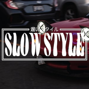May include: A red car with the words "Slow Style" in white text on a black background. The car has a black bumper and a black grill. The car is in focus, and the background is blurred.