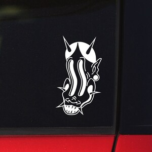 May include: A white vinyl sticker featuring a stylized devil face with sharp horns, a wide grin, and elongated eyes. The sticker is applied to a black surface, likely a car window, with a red car body visible in the background.
