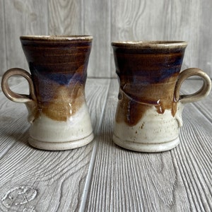 以下が含まれることがあります： 茶色の釉薬がかかった、茶色と白のセラミック製のマグカップが2つ。マグカップは、幅広の底と狭くなった上部というユニークな形をしています。マグカップは木製の表面の上に置かれています。