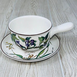 May include: A white ceramic soup bowl with a floral design and a handle. The bowl is on a matching saucer with a floral design. The bowl and saucer are on a light wood surface.