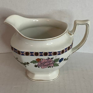 Puede incluir: Crema de cerámica blanca con un diseño floral en rosa, azul y dorado. La crema tiene una base cuadrada y un asa.