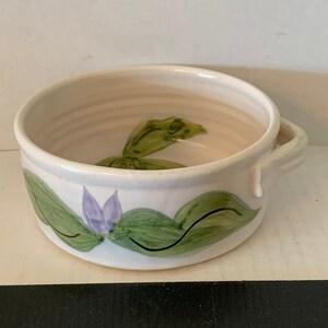May include: A white ceramic bowl with a green and purple floral design. The bowl has a handle on one side.