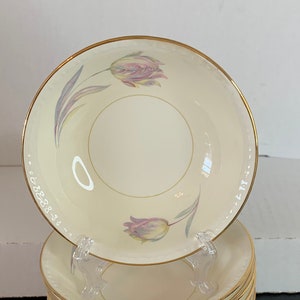May include: A stack of 12 white ceramic bowls with a gold rim and a floral design. The bowls are stacked on top of each other, with the top bowl showing a pink tulip design.