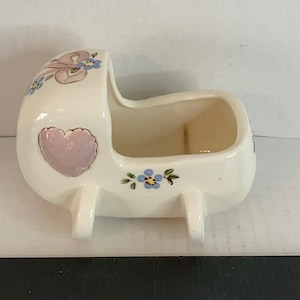 Puede incluir: Un macetero de cerámica blanca en forma de cochecito de bebé con detalles florales rosas y azules. El cochecito tiene un corazón rosa en el lateral y flores azules en la parte inferior.