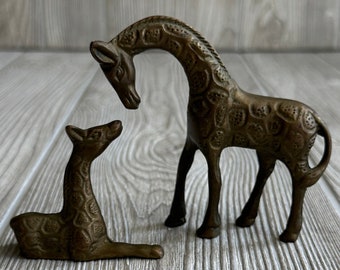 May include: Two brass giraffe figurines, one standing and one lying down, with a textured surface. The standing giraffe has its head bent down, as if it is looking at the lying giraffe.
