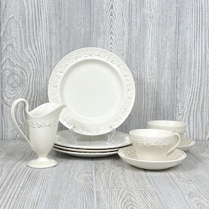 Puede incluir: Un juego de té de porcelana color crema con una jarra de crema, platos, platillos y tazas de té. El juego tiene un diseño floral en relieve alrededor de los bordes de los platos, tazas y jarra de crema.