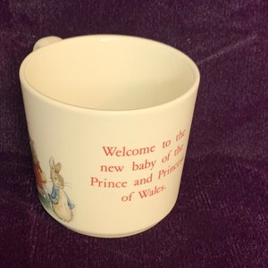 Puede incluir: Taza de cerámica blanca con una ilustración rosa y azul de Peter Rabbit y Benjamin Bunny. La taza tiene el texto "Welcome to the new baby of the Prince and Princess of Wales."