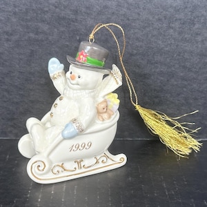 May include: A white porcelain snowman ornament riding in a gold sleigh with a blue hat and a red flower. The ornament is dated 1999.