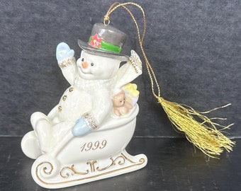 May include: A white porcelain snowman ornament riding in a gold sleigh with a blue hat and a red flower. The ornament is dated 1999.