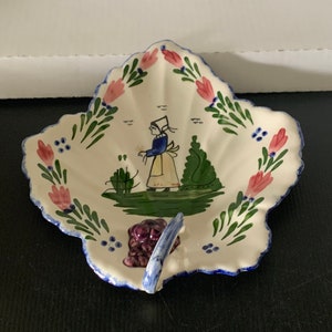May include: A white ceramic leaf-shaped dish with a blue and green painted design. The dish features a woman in a blue dress and white apron, holding a basket of grapes. The dish has a purple handle.