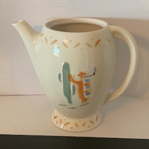 May include: A white ceramic teapot with a brown, green, and blue cactus and person design. The teapot has a handle and a spout.