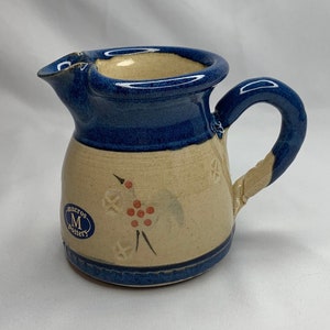 Puede incluir: Una pequeña jarra de cerámica azul y crema con un diseño floral y un logotipo azul y blanco que dice "Meredith Pottery".