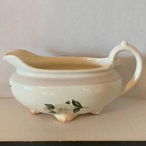 May include: White ceramic creamer with a floral design and three feet. The creamer has a light pink rim and a handle.