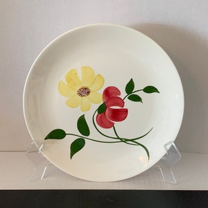 May include: A white ceramic plate with a floral design. The plate features a yellow flower with a red center and a red flower with a yellow center. The flowers are painted on the plate with green leaves.