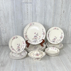 Puede incluir: Un juego de vajilla de porcelana vintage con estampado floral, que incluye platos, cuencos y una fuente para servir. Los platos son de color crema con diseños florales rosas y morados.