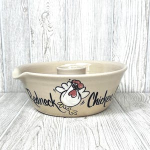 May include: Tan ceramic bowl with a spout and a central cylinder. The bowl features a cartoon chicken graphic with the words "Redneck Chicken" in black script. The bowl is set against a light wood-grain background.