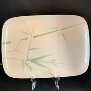 May include: A rectangular, off-white plate with rounded corners. The plate features a delicate bamboo design in shades of green. The plate is displayed on a clear plastic stand against a dark background.