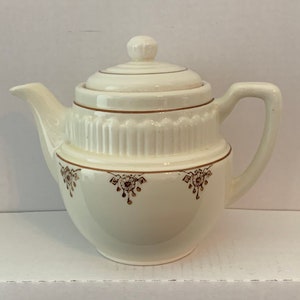 May include: Cream-colored ceramic teapot with a lid and handle. The teapot features brown floral patterns and trim. The lid has a small knob. The teapot is on a white surface against a white background. A classic tea set item.