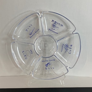 Puede incluir: Un plato de Seder de plástico transparente con seis secciones. Cada sección está etiquetada con texto hebreo y una imagen que representa el artículo del plato de Seder. Las secciones están etiquetadas: Hueso de la pierna, Haroseth, Huevo, Hierbas amargas, Perejil y Karpas.