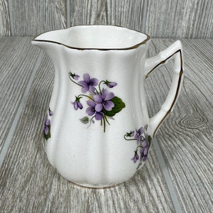 May include: A white ceramic creamer with a gold trim and a floral design featuring purple violets.
