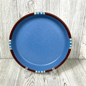以下が含まれることがあります： 丸い青いセラミックプレート。濃い茶色の縁があり、白とターコイズのストライプが特徴です。透明なスタンドに載せられ、明るい木製の背景に配置されています。