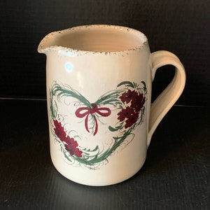 以下が含まれることがあります： 緑と赤の花柄が描かれた白い陶器のピッチャー。ピッチャーはハート型で、中央に赤いリボンが結ばれています。