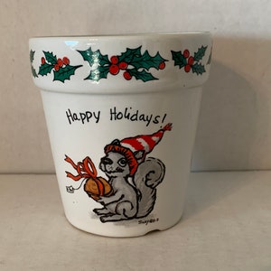 May include: White ceramic planter with a green holly border and a squirrel wearing a red and white striped hat holding a nut. The planter says "Happy Holidays!"
