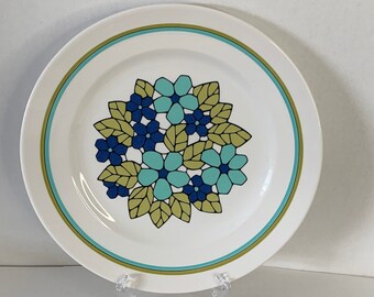 May include: A white ceramic dinner plate with a green, blue, and yellow floral design. The plate has a thin green and blue border.