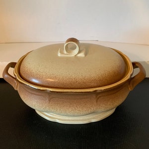 Puede incluir: Una fuente de cerámica cubierta con un diseño moteado beige y marrón. La fuente tiene dos asas y una tapa con un pequeño asa redonda. La base es de color beige claro, y el borde es de color marrón oscuro.
