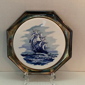 May include: A white ceramic coaster with a blue and white image of a sailing ship on a stormy sea. The coaster is framed in a silver octagon.