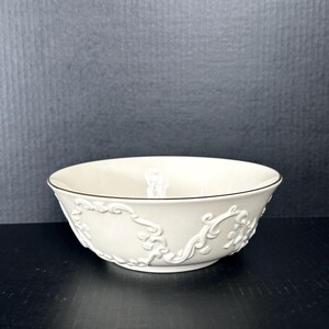 May include: A white ceramic bowl with an embossed floral design. The bowl has a black rim and is perfect for serving snacks or salads.
